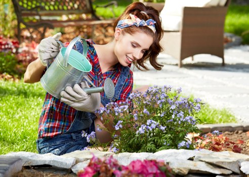 A young woman with light skin, wearing a plaid shirt, denim overalls, gardening gloves, and a colorful headband, is tending to a flower bed in a well-maintained garden outside a residential property. She is smiling as she waters a dense cluster of small purple and light blue flowers with a metal watering can. The garden features a variety of plants, including a neatly edged flower bed and a lawn with lush green grass. In the background, there is a paved patio area with outdoor furniture, including a table and chairs, alongside a backdrop of trees and shrubs creating a natural, inviting outdoor setting. The weather appears sunny with bright natural light illuminating the scene, indicating a pleasant day suitable for outdoor gardening work. This image reflects professional gardening care typical of services provided by landscapers in Brixton, emphasizing vibrant plant growth and tidy landscape maintenance.
