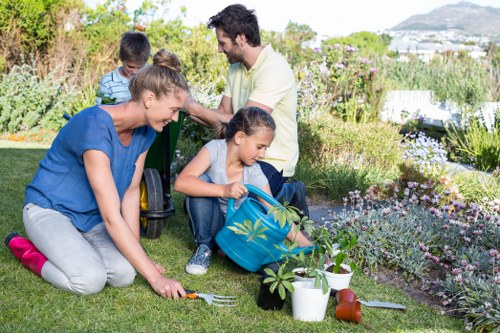 A family engaged in gardening activities in a well-maintained backyard, featuring a lush green lawn with a variety of flowering plants and shrubs along the flower beds. The foreground shows a woman with blonde hair kneeling on the grass, planting or tending to a small tree or shrub, with gardening tools and potted plants nearby. Two children, a girl and a younger boy, are sitting on the grass; the girl is holding a small rake or gardening tool and a blue backpack, while the boy is watching. In the background, a man with dark hair, wearing a light-colored shirt, is standing or crouching near the plants, possibly assisting or supervising. The garden includes a paved pathway or patio area on one side, and the scene is bathed in natural daylight, suggesting a pleasant, sunny day in Brixton, with a scenic view of distant houses and greenery beyond the garden. This outdoor setting showcases a well-kept garden space ideal for family gardening and outdoor activities, aligning with professional landscaping and gardening services such as those offered by Landscapers Brixton.