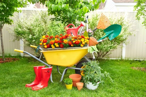 A vibrant garden scene in a backyard with a well-maintained green lawn and a variety of garden elements. A yellow wheelbarrow filled with bright red and orange flowers sits centrally, surrounded by gardening tools including a red watering can, a pair of red rubber boots, and several small terracotta pots placed on the grass. Behind the wheelbarrow, there is a leafy tree with green foliage providing partial shade, and a white wooden fence borders the garden space, ensuring privacy. To the right of the wheelbarrow, a white potted plant with lush green leaves adds to the garden’s sense of natural abundance. The overall environment appears bright and sunny, suggesting warm weather ideal for outdoor gardening activities. This outdoor space, typical of residential gardens in Brixton, showcases neatly arranged plants and gardening equipment that reflect a well-kept and inviting outdoor area, consistent with services offered by Landscapers Brixton for garden landscaping, lawn care, and outdoor maintenance.