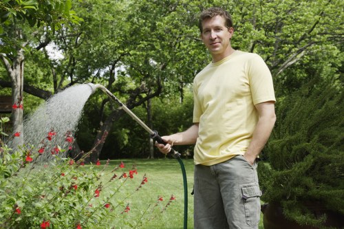 A man stands in a well-maintained garden in Brixton, holding a garden hose and watering a flower bed with red blossoms and green foliage. The garden features a lush, green lawn with dense trees and shrubs in the background, providing shade and natural beauty. The flower bed borders are clearly defined, and the garden surface area appears tidy and healthy. The man is casually dressed in a light yellow t-shirt and grey cargo shorts, enjoying outdoor maintenance on a bright, slightly overcast day. Surrounding him are natural textures of soil, grass, and foliage, contributing to a vibrant outdoor environment. This scene exemplifies typical garden care activities which a professional landscapersbrixton.co.uk gardening service like Landscapers Brixton could support, ensuring gardens are lush, organised, and visually appealing.