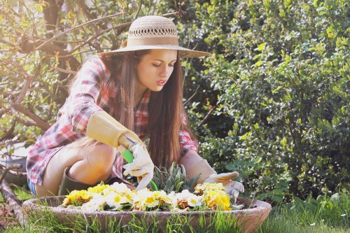 A young woman wearing a straw sun hat, plaid shirt, and gardening gloves is kneeling in a lush garden with dense green foliage in the background. She is carefully tending to a flower bed filled with a mixture of yellow, white, and purple blooming flowers. The garden features a well-maintained lawn with vibrant grass surrounding the flower bed, and the sunlight creates soft highlights on her hair and the foliage. The scene suggests a serene outdoor space typical of residential gardens in Brixton, with natural tones and an emphasis on healthy plant growth, reflecting professional gardening and landscaping practices by Landscapers Brixton. This image captures the quiet, attentive moment of gardening activity suitable for a gardening service webpage focusing on flower bed maintenance and outdoor garden enhancement.