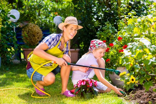 A woman and a young girl are gardening together in a lush backyard garden, with the woman wearing a straw hat, plaid shirt, and yellow apron, smiling as she assists the girl. The girl, dressed in a white outfit with a floral hat, is crouched down, carefully tending to a flower bed bordered by colorful blooming plants, including red, yellow, and pink flowers. The garden features a well-maintained grass lawn, flower beds with rich soil, and background foliage of trees and hedges, providing a vibrant and natural outdoor setting. Decorative lanterns hang among the greenery, indicating a thoughtfully landscaped backyard. The woman holds a yellow garden hose, suggesting ongoing watering or gardening maintenance. The scene is naturally lit, with bright sunlight casting soft shadows, highlighting the healthy plants and lush environment. This outdoor space is typical of a residential garden in Brixton, designed for family gardening activities and outdoor enjoyment, supported by local gardening services such as Landscapers Brixton.